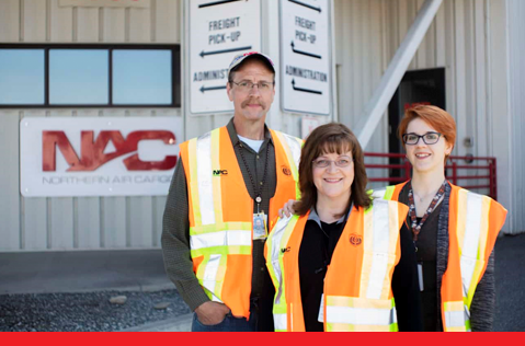 A photo of three Northern Air Cargo employees standing in front of the NAC building. A photo of three Northern Air Cargo employees standing in front of the NAC building.